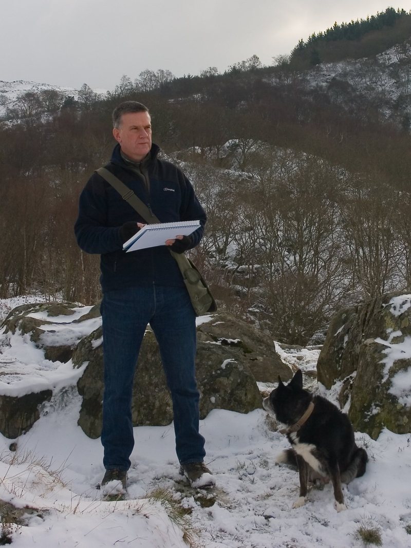 Chris Hull standing on a snowy hillside with a sketchbook, his dog sitting beside him